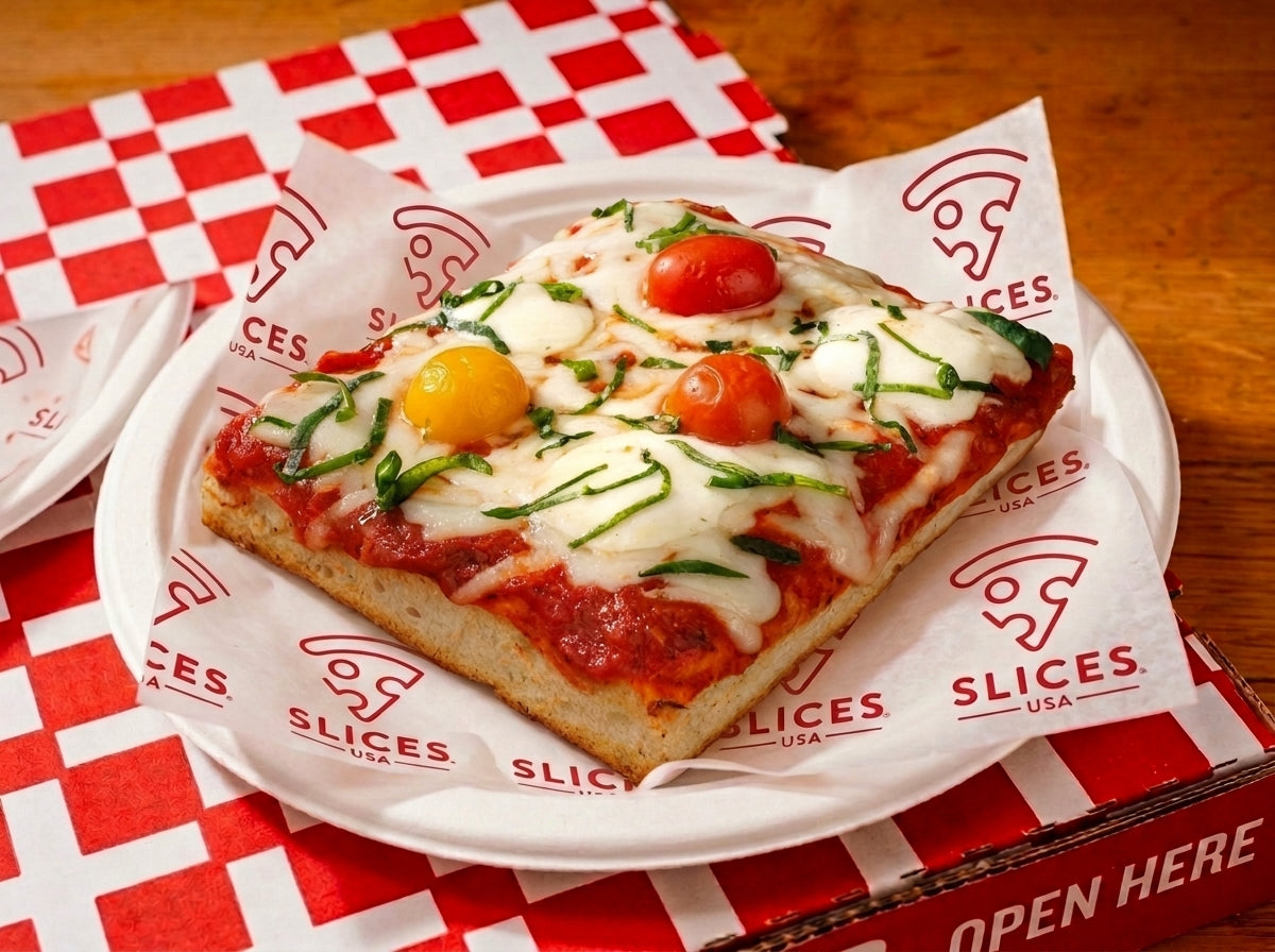 A square slice of pizza on a white plate, topped with melted cheese, green basil, and cherry tomatoes, served on branded paper over a red-and-white checkered table setting.