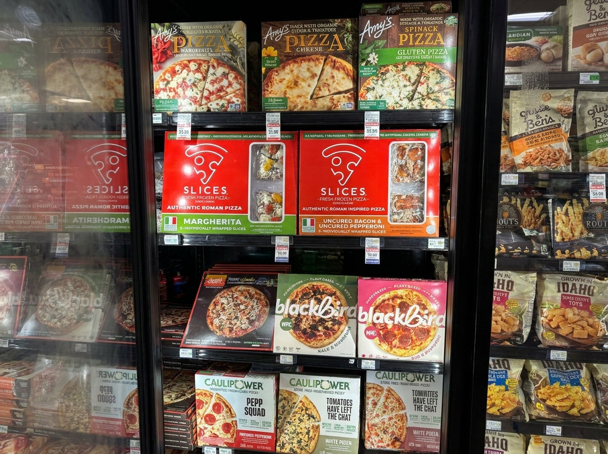 A grocery store freezer display filled with packaged frozen foods, prominently showing multiple pizza boxes along with other meals and snacks on the shelves.