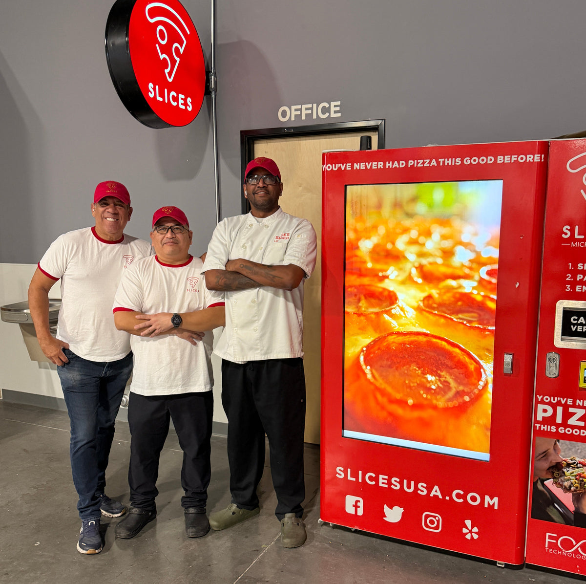 Three men in white and red Slices pizza uniforms stand next to a large red Slices vending machine in an office-like indoor space. A red round Slices sign hangs on the wall above them.
