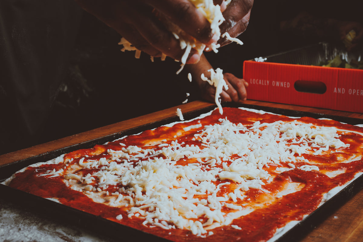 A close-up of a pizza dough sheet topped with red tomato sauce and being sprinkled with shredded white cheese by hands, with a red branded delivery box in the background.