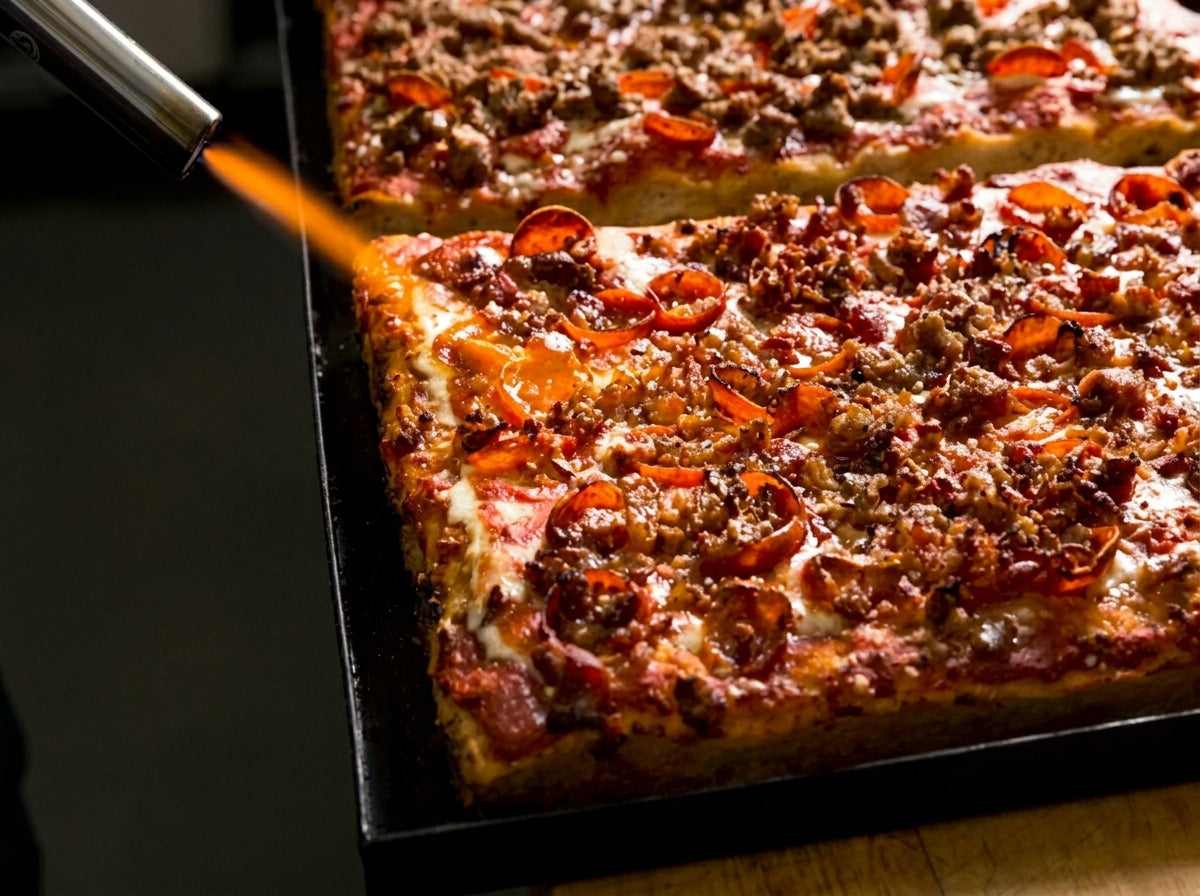 A close-up of a square pizza topped with sausage crumbles, melted cheese, and sliced red peppers, being toasted with a torch on a dark baking pan.